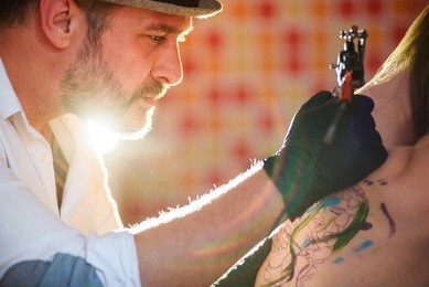 portrait of professional creative tattoo master wearing hat and gloves making tattoo on a young girl's back in tattoo salon. tattoo making process with machine gun.