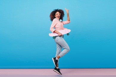 indoor full-length portrait of confident african girl in pink shirt holding skateboard. enthusiastic black woman with curly hairstyle posing in studio with blue interior.