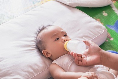 happy mother holding and feeding milk for her asian newborn baby boy from plastic bottle at home. breastfeeding or early childhood development concept