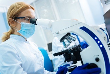 making medical discovery. serious blond young researcher wearing a uniform and a medical face mask and gloves and glasses and looking in the microscope while being in the lab