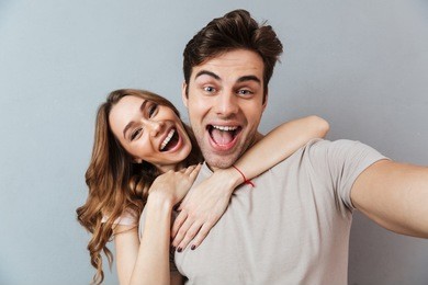 portrait of an excited young couple hugging while standing and taking a selfie over gray wall
