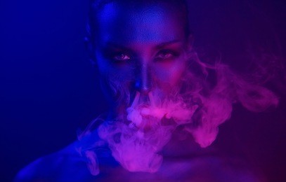 neon night. halloween vape party. beautiful sexy young vamp woman with glamorous mystical makeup vaping in nightclub (exhaling smoke). girl smoking in club. blue mystic smoke 