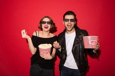 laughing punk couple in eyeglasses eating popcorn and looking at the camera over red background