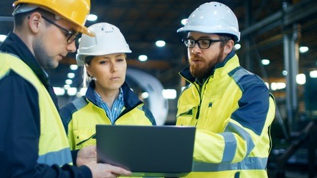 male and female industrial engineers talk with factory worker while using laptop. they work at the heavy industry manufacturing facility.