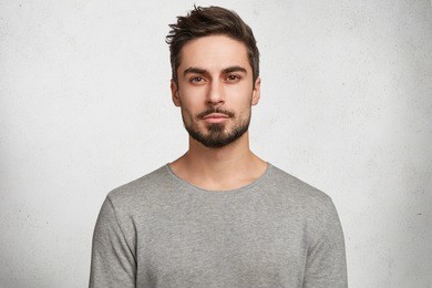 isolated shot of young handsome male with beard, mustache and trendy hairdo, wears casual grey sweater, has serious expression as listens to interlocutor, poses in studio against white background