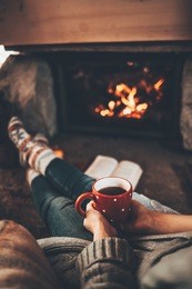 feet in woollen socks by the christmas fireplace. woman relaxes by warm fire with a cup of hot drink and warming up her feet in woollen socks. cozy atmosphere. winter and christmas holidays concept.