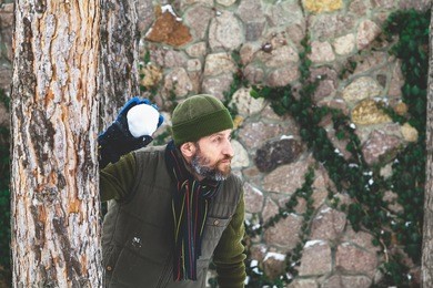 playing snowballs between trees. a man with a beard in a warm waistcoat and hat is throwing snowballs between the trunks of pine trees during a frosty day
