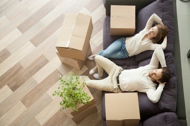 couple resting on couch after moving in, man and woman relaxing on sofa just moved into apartment with cardboard boxes on floor, happy satisfied homeowners enjoying first day in new home, top view