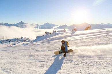 a snowboarder in a ski mask and a backpack is riding on a snow-covered slope leaving behind a snow powder against the blue sky and the setting sun