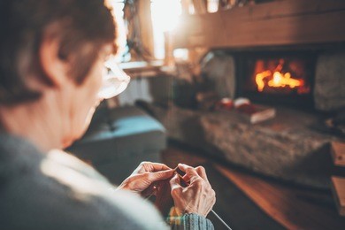 senior woman hands knitting by the fireplace. unrecognisable grandmother relaxes by warm fire making handmade gifts for her family. cozy atmosphere. winter and christmas holidays concept.