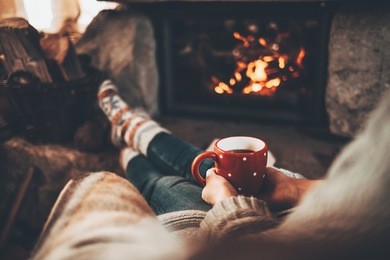 woman in warm knitted cardigan sweater by fireplace. woman relaxes by warm fire with a cup of hot drink and warming up her feet in woollen socks. cozy atmosphere. winter and christmas holidays concept