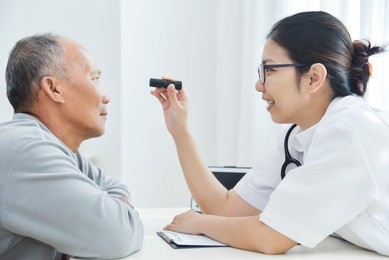 young asian female doctor wear glasses checking senior man patient eyes with flashlight in medical office.