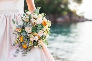 bride holds wedding bouquet from roses and stands on background of sea and beach after wedding ceremony. bride is dressed in cream wedding dress.