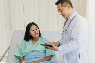 cheerful asian doctor talking with young woman patient for monitoring and check up after surgery. doctor giving advice in the touchscreen to women to take care the lesion after out of hospital.