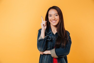 happy asian woman in denim jacket pointing up and looking at the camera over yellow background