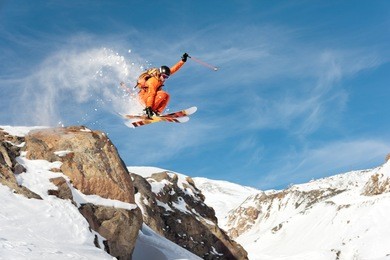 a professional skier makes a jump-drop from a high cliff against a blue sky leaving a trail of snow powder in the mountains