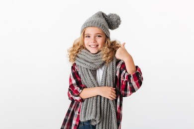portrait of a smiling little girl dressed in winter hat and scarf showing thumbs up isolated over white background