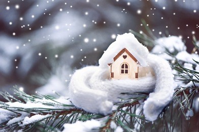 christmas toy house is wrapped in a warm scarf, it's snowing.on a natural natural background of a real fir in the snow, toned. concept of winter, christmas, new year,
 warm, cozy, loving, protecting t