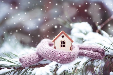 christmas toy house is wrapped in a warm scarf, it's snowing.on a natural natural background of a real fir in the snow, toned. concept of winter, christmas, new year,
 warm, cozy, loving, protecting 