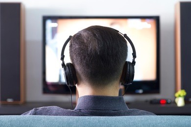 back side of an asian man wearing black heaphones in front of blurry out-of-focus television and home entertainment system background