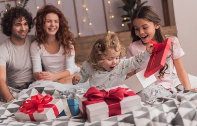 it's christmas time! happy family sitting in bed, parents are smiling while their daughters are opening the presents