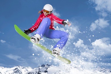 snowboarder in red jacket and lilac trousers jumps on her green snowboard through the air in front of a mountain panorama and winter landscape
