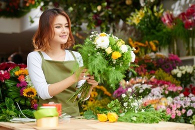 portrait of young asian florist wearing apron standing at counter and focused on making bouquet for client, interior of flower shop on background