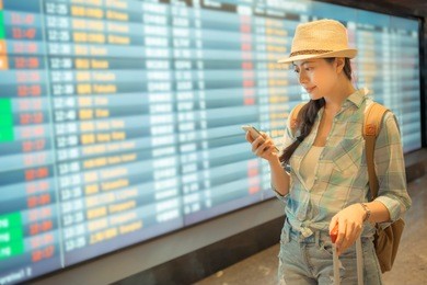 young asian woman with phone in her hand, tickets in pocket of shoulder bag checking flight timetable in international airport. travel concept.