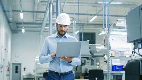 chief engineer in the hard hat walks through light modern factory while holding laptop. successful, handsome man in modern industrial environment.