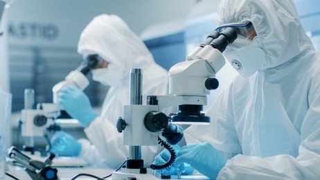 two engineers/ scientists/ technicians in sterile cleanroom suits use microscopes for component adjustment and research. they work in an electronic components manufacturing factory.