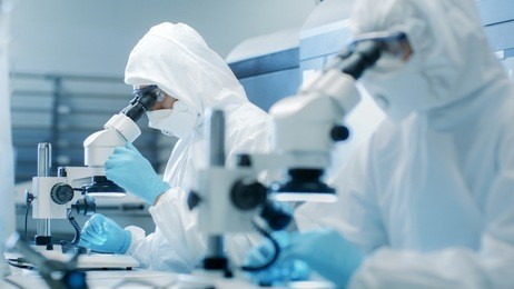 two engineers in sterile cleanroom suits  control manufacturing machinery work and use microscopes for component adjustment and research. they work in an electronic components manufacturing factory.