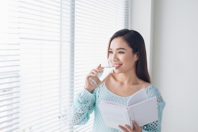 beautiful asian girl drinking water and reading book near window at home