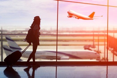 young woman is standing near window at the airport and watching plane before departure. she is standing and carrying luggage.travel concept .