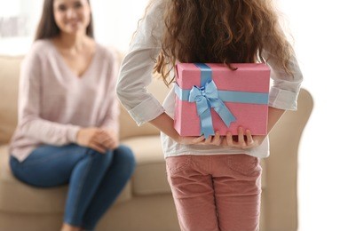 little girl hiding gift box for mom behind her back indoors. mother's day celebration