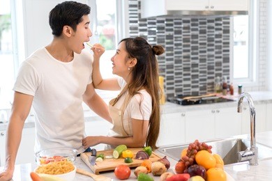 couple together in kitchen room, young asian woman holding vegetables to man eating each for cooking dinner healthy food menu at home couple together romantic 
