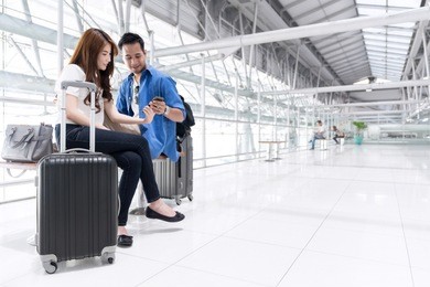 young happy teenage asian couple check in by mobile and waiting in airport terminal. couple sitting and using smartphone together, air travel or holiday vacation concept.