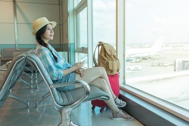 woman passenger traveler in airport waiting for air travel holding smart phone, smiling sitting with travel suitcase trolley, in waiting hall of departure lounge in airport.
