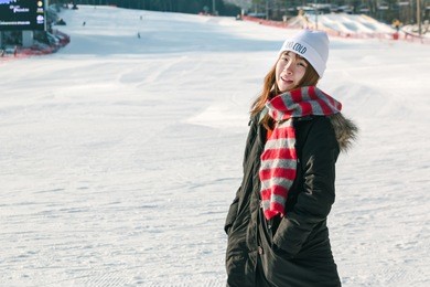 winter asian woman smiling in snow. beautiful young asian woman enjoying the snow in skii resort dressed in a black coat, cheerful red strip winter scarf and white hat. happy winter holiday.