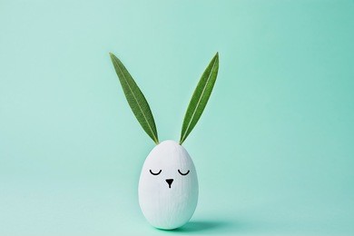 decorative white painted easter egg bunny with drawn cute kawaii face. green leaves as ears. pastel turquoise background. spring holiday crafts kids concept. greeting card poster banner. copy space 