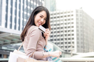 beautiful young smiling asian woman carrying shopping bags in her arms presenting credit card that just used for making payment