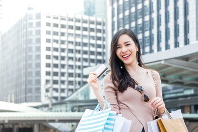 beautiful young smiling asian woman carrying shopping bags in her arms presenting credit card that just used for making payment