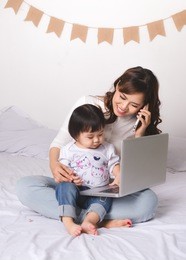 asian lady in classic suit is talking on the mobile phone and working on laptop at home with her baby girl.