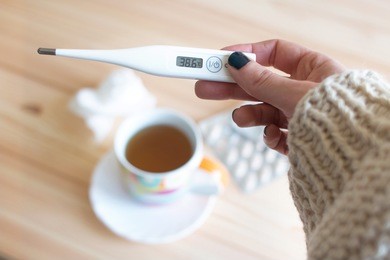 close up of a hand holding a digital thermometer over a cup of tea , a papper tissue and a flu medicaments. cold and flu concepts
