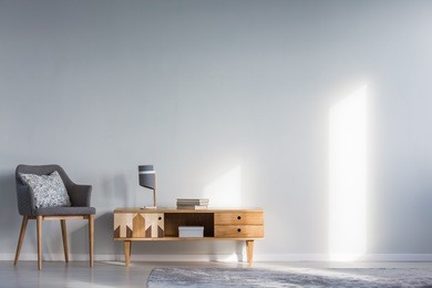 simple living room interior with patterned cushion lying on a gray armchair, next to a wooden cupboard with drawers