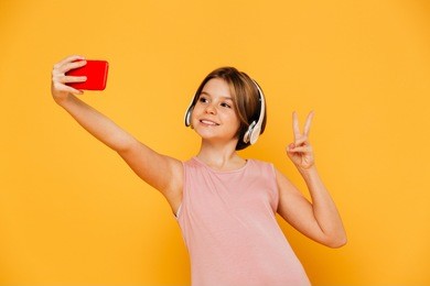 pretty modern girl in headphones smiling and making selfie on smartphone isolated over yellow