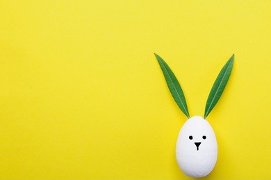 decorative white painted easter egg bunny with drawn cute kawaii face. green leaves as ears. pastel yellow background. spring holiday crafts kids concept. greeting card poster banner. copy space