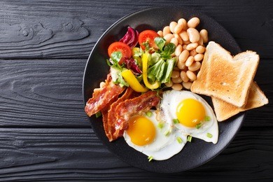 delicious fried eggs, bacon, beans, toast and fresh vegetable salad on a plate on the table. horizontal top view from above