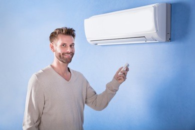 happy young man holding remote standing in front of air conditioner