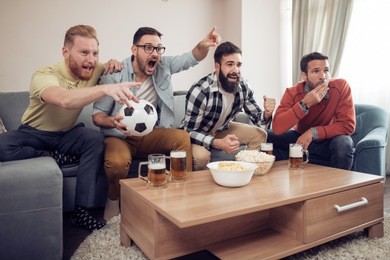 young football fans are drinking beer,cheering and smiling while watching football game at home.