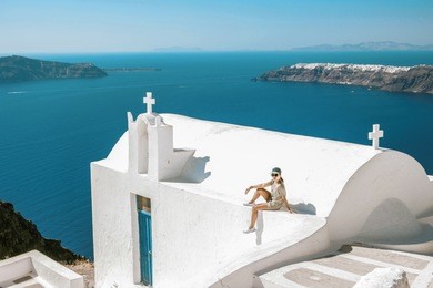 santorini of a typical greek church overlooking the rock of skaros with the town of oia and the volcano in the background, young woman at a greek white church looking over the ocean at fira santorini